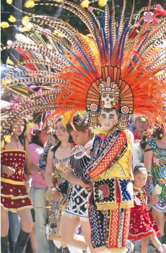 Danzante con enorme penacho de plumas y máscara de calavera prehispánica, entre other participantes y público.
