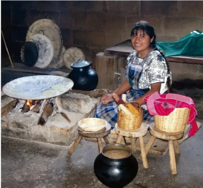 Mujer indígena sonríe sentada junto a un fogón de leña; sobre el fuego hay un comal con tortillas y alrededor ollas de barro, canastas tejidas y utensilios tradicionales de cocina.
