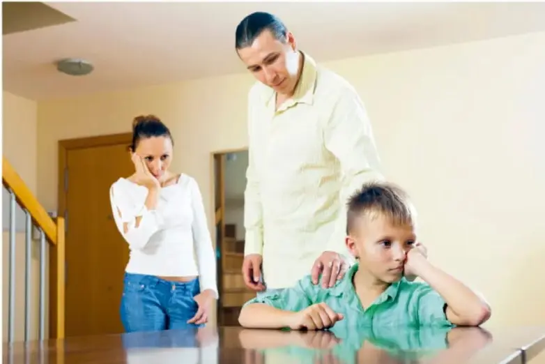 Fotografía de una familia en el interior de una casa: un niño sentado con la mano en la mejilla y expresión de tristeza o aburrimiento, mientras una mujer y un hombre de pie lo observan con preocupación.