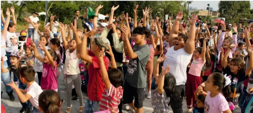 Niñas, niños y algunas personas adultas al aire libre con los brazos levantados y expresiones de alegría, celebrando durante un evento comunitario.