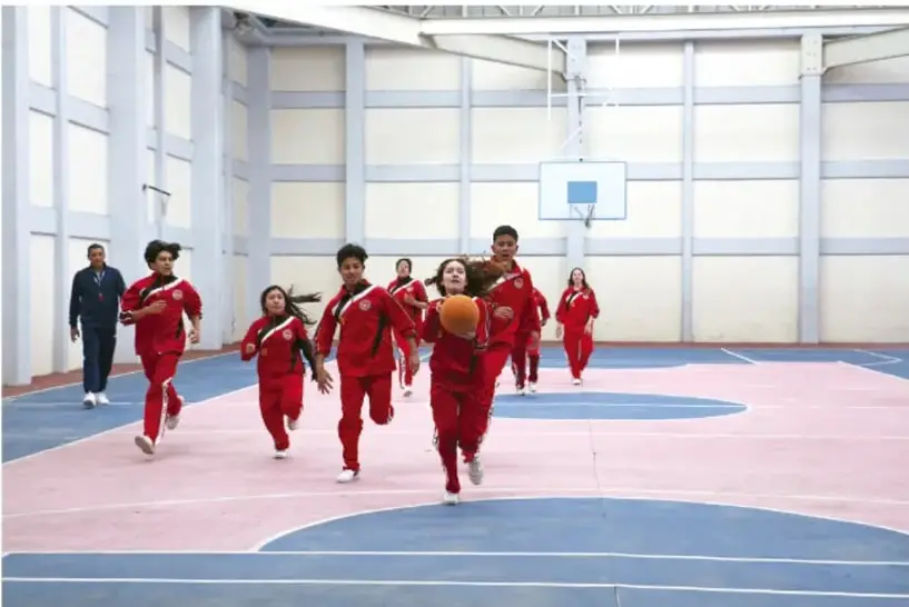 Fotografía de un grupo de estudiantes con uniforme rojo corriendo en un patio escolar cubierto con cancha de básquetbol; una estudiante conduce el balón y un docente los acompaña al costado.