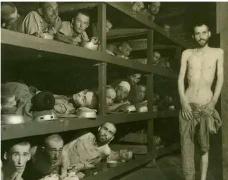Fotografía histórica en blanco y negro de prisioneros en un campo de concentración nazi, apiñados en literas de madera; un prisionero demacrado y semidesnudo está de pie en el extremo derecho.