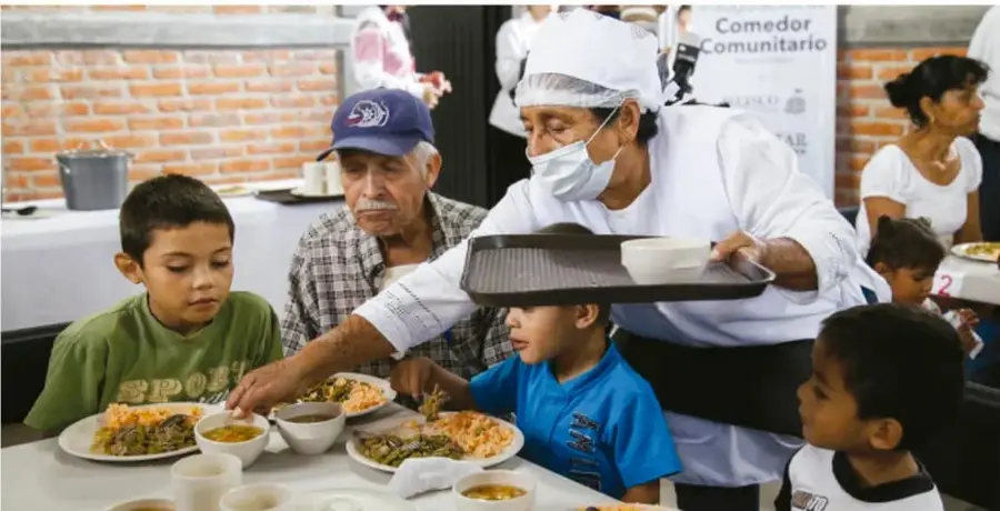 Personas de una comunidad recibiendo alimentos en un comedor comunitario, atendidas por una cocinera con cubrebocas y una persona mayor.