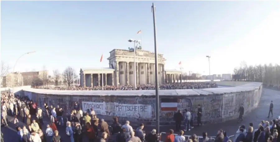 Fotografía de la Puerta de Brandeburgo con una multitud reunida frente a un tramo del Muro de Berlín cubierto de grafitis, en el contexto de su caída en 1989.