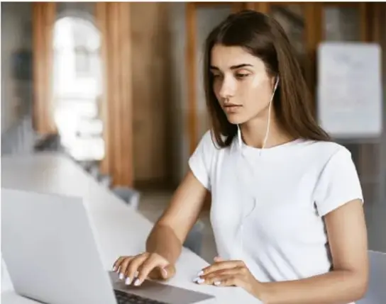 Fotografía de una joven con audífonos trabajando frente a una laptop en un espacio de trabajo compartido, ilustrando el uso de la tecnología.