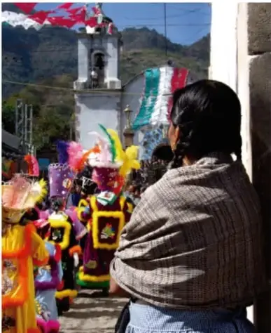 Fotografía de una persona de espaldas con manta tejida observando un desfile o festividad con figuras coloridas, globos y una iglesia al fondo, en un paisaje montañoso.