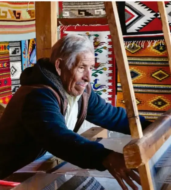 Fotografía de un artesano indígena mayor trabajando en un telar de madera, con textiles y tapetes de colores y figuras geométricas colgados al fondo.