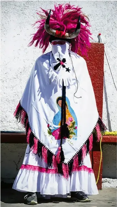 Fotografía de una persona con traje tradicional de Baja California: falda blanca con flecos rosas y negros, capa bordada con imagen religiosa, máscara con penacho de plumas rosas y cuernos. Crédito: Sandra Angélica Martínez Cruz, Baja California.