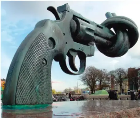 Escultura de bronce de un revólver con el cañón anudado, conocida como 'Non Violence', símbolo de la no violencia. Al fondo se observan árboles y un espacio público.
