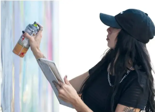 Fotografía de una persona joven con gorra negra pintando un mural con aerosol; sostiene una libreta o cuaderno en la mano izquierda.