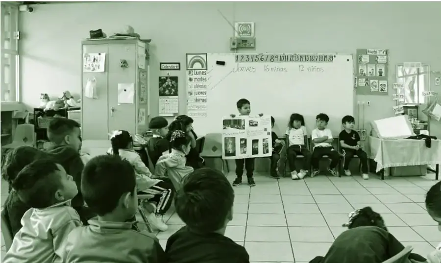 Fotografía de un aula de preescolar: una niña o niño muestra un trabajo en cartulina frente a sus compañeros sentados en sillas y en el suelo durante una asamblea; al fondo hay un pizarrón con la secuencia numérica 1 a 21, días de la semana (lunes, martes, miércoles, jueves, viernes) y carteles del aula.