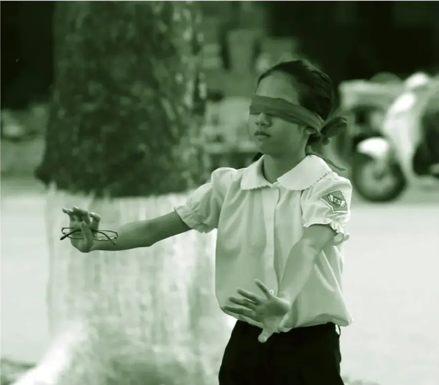 Fotografía en tonos verdes de una niña con los ojos vendados con una tela, brazos extendidos y un objeto en la mano, jugando gallinita ciega al aire libre; al fondo un árbol y una persona en bicicleta.