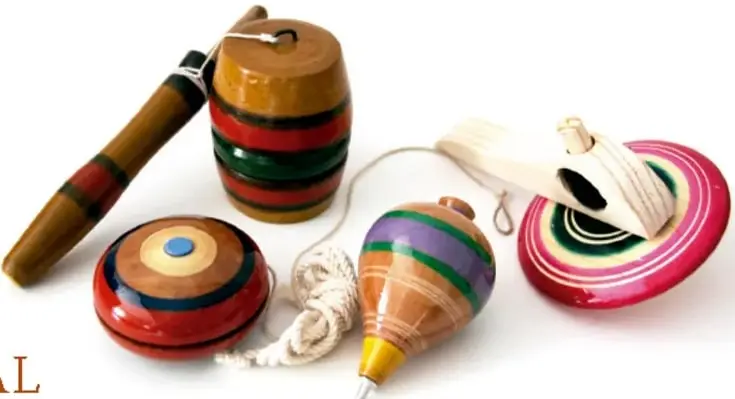 Fotografía de juguetes tradicionales mexicanos: un balero de madera con franjas de colores, un trompo pintado, un yoyo rojo con azul y un yoyo rosa con blanco, todos sobre fondo blanco.