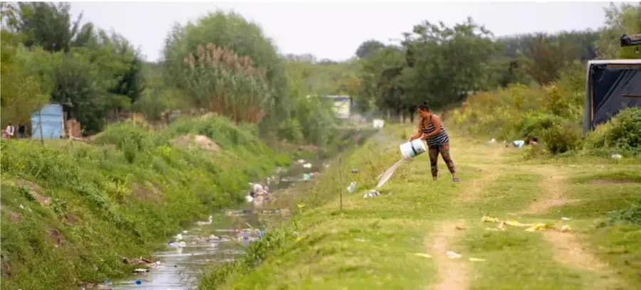 Fotografía de un canal o escurrimiento con basura a los lados, donde una persona vierte el contenido de un balde cerca del agua, ilustrando una situación de riesgo ambiental y de salud en el entorno.