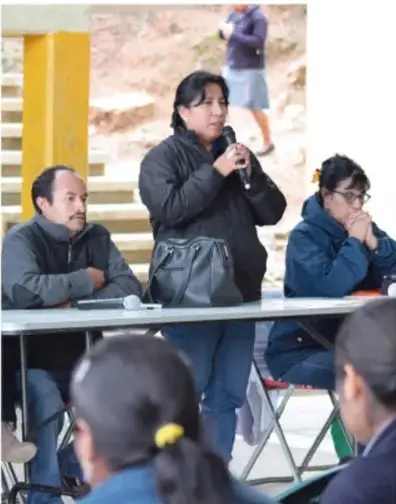 Fotografía de una reunión comunitaria al aire libre: una mujer de pie habla por micrófono mientras varias personas sentadas escuchan y toman notas.