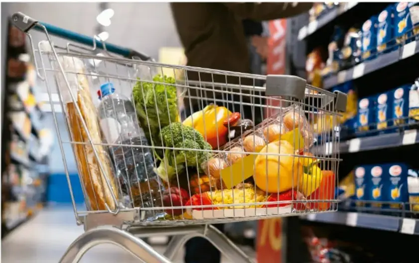 Carrito de supermercado con alimentos saludables: pan, agua embotellada, brócoli, jitomates, zanahorias, manzanas y otras frutas y verduras; al fondo, estantes de una tienda.