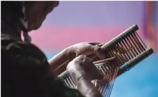 Fotografía de las manos de una artesana trabajando en un telar de cintura, con hilos de colores tensados; fondo con tonos azul y rosa.