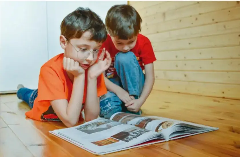 Fotografía de dos niños sentados en el suelo de madera leyendo un libro abierto; uno usa lentes y playera naranja, el otro playera roja.
