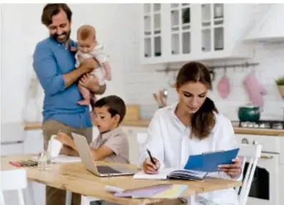 Familia en la cocina: un hombre carga a un bebé, una mujer escribe en una libreta y un niño usa una laptop.