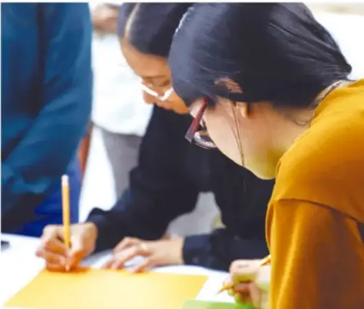 Fotografía de estudiantes trabajando con lápiz y hojas de colores en equipo, ilustrando el trabajo colaborativo.