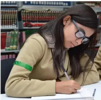 Fotografía de una estudiante (Diana) con suéter beige y brazalete verde, escribiendo su diario en un banco frente a una librería.