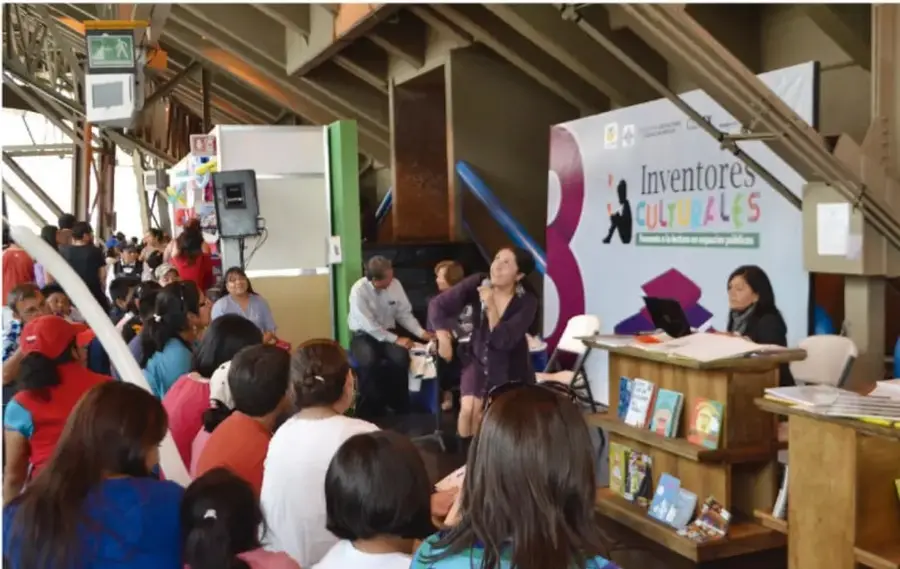 Fotografía de una presentación cultural en un espacio público techado; una mujer habla frente a un público sentado y hay un cartel que dice «Inventores culturales» junto a una mesa con libros.