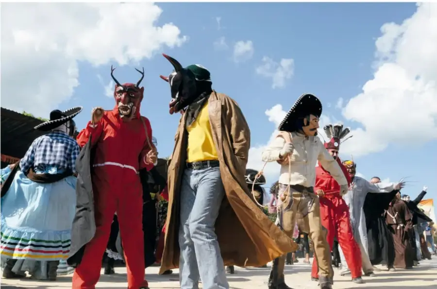 Comparsa de Días de Muertos al aire libre: danzantes disfrazados de diablos de traje rojo y de personaje con máscara, sombrero y capa, junto a otros participantes con vestimenta regional, bajo cielo con nubes.
