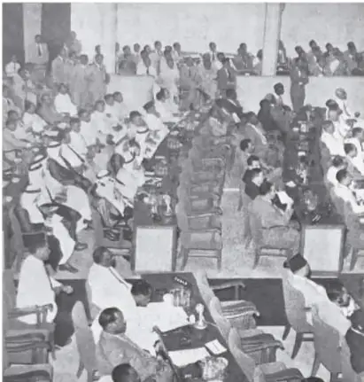 Fotografía en blanco y negro de una asamblea numerosa, posiblemente la Conferencia de Bandung, con delegados de pie y sentados.