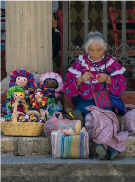 Mujer indígena anciana tejiendo, sentada junto a una cesta con muñecas de trapo de colores y textiles; fotografía que ilustra la permanencia de las culturas indígenas.