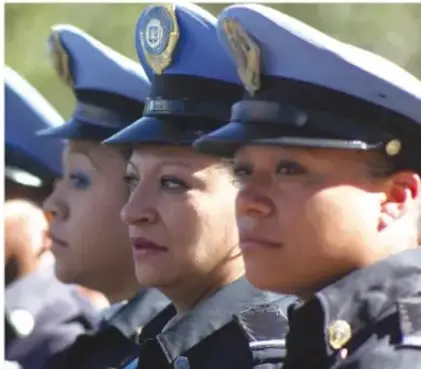 Fotografía de tres funcionarias policiales en formación con uniforme azul oscuro, gorras de plato y rodaje, en fila con mirada al frente (mujeres en la función pública de seguridad).