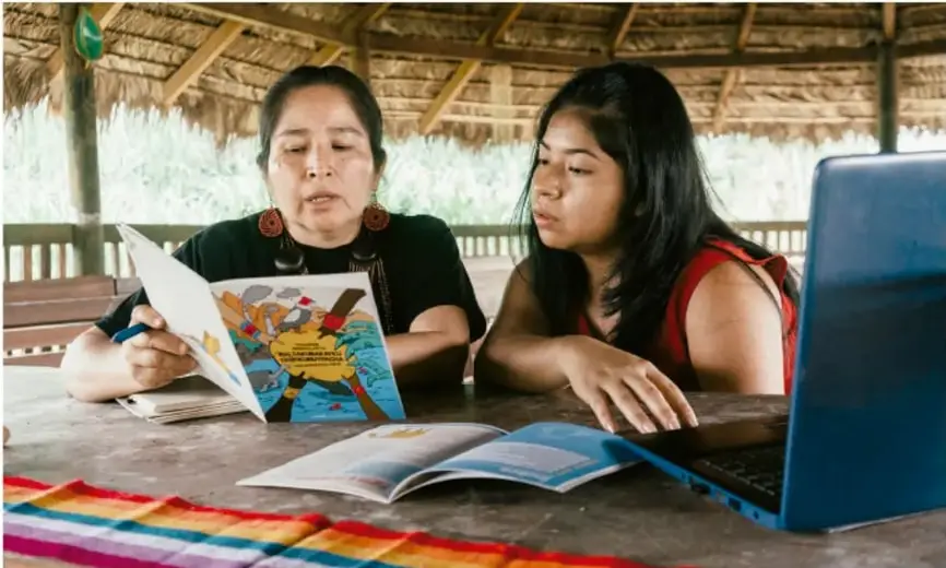 Fotografía de dos mujeres (una adulta con aretes y una joven) sentadas bajo un rancho de palapa, revisando juntas un tríptico impreso con lámina de salud e identidad comunitaria, con un laptop azul sobre la mesa y un textiles de colores en primer plano; representan la revisión colaborativa de los productos del proyecto.
