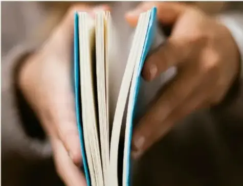 Fotografía de un par de manos sosteniendo un libro abierto con las páginas ligeramente abanico, sobre un fondo borroso.