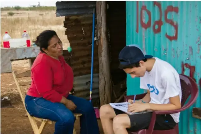 Fotografía: una estudiante con gorra toma notas en un cuaderno mientras conversa con una mujer sentada frente a una vivienda sencilla de lámina, en un entorno rural; ilustra la investigación de campo o entrevista dentro del proyecto académico.