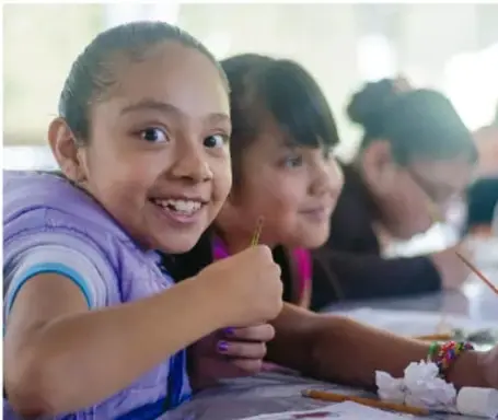 Fotografía de niñas sonriendo en un aula escolar, escribiendo o dibujando, representando el bienestar integral.