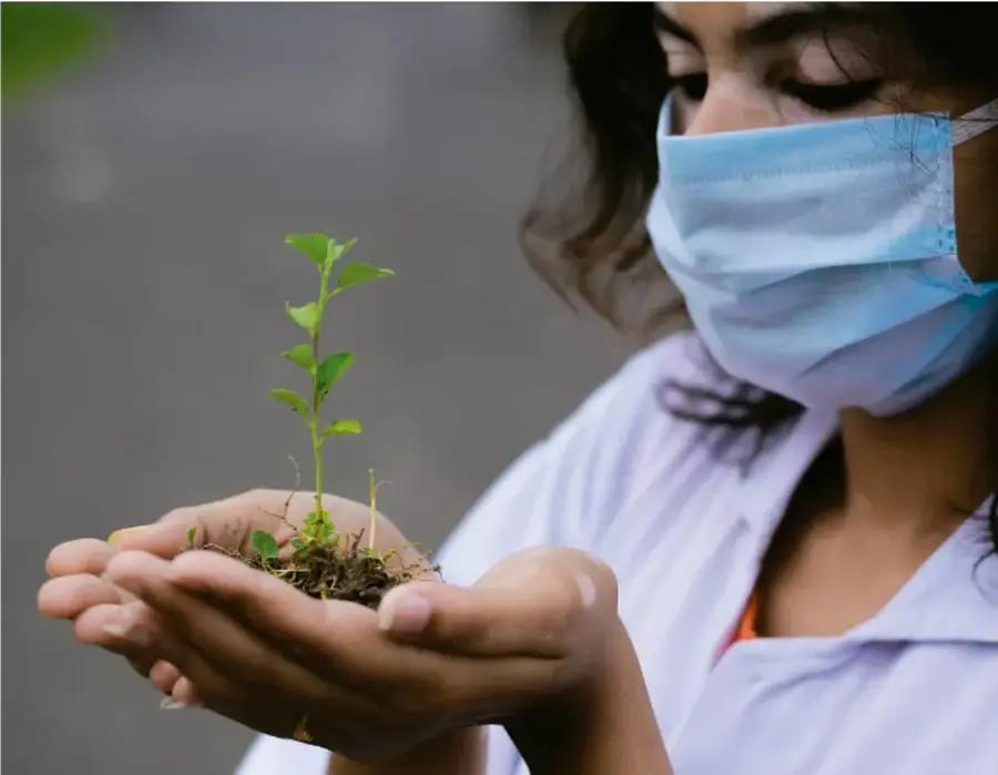 Fotografía de una estudiante con mascarilla sanitaria y bata escolar sosteniendo entre sus manos una planta joven con tierra, símbolo de cuidado ambiental.