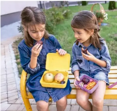 Fotografía de dos niñas sentadas en una banca del patio escolar. Una sostiene una lonchera amarilla con fruta y la otra come de un recipiente morado. Al fondo hay pasto y una canasta.