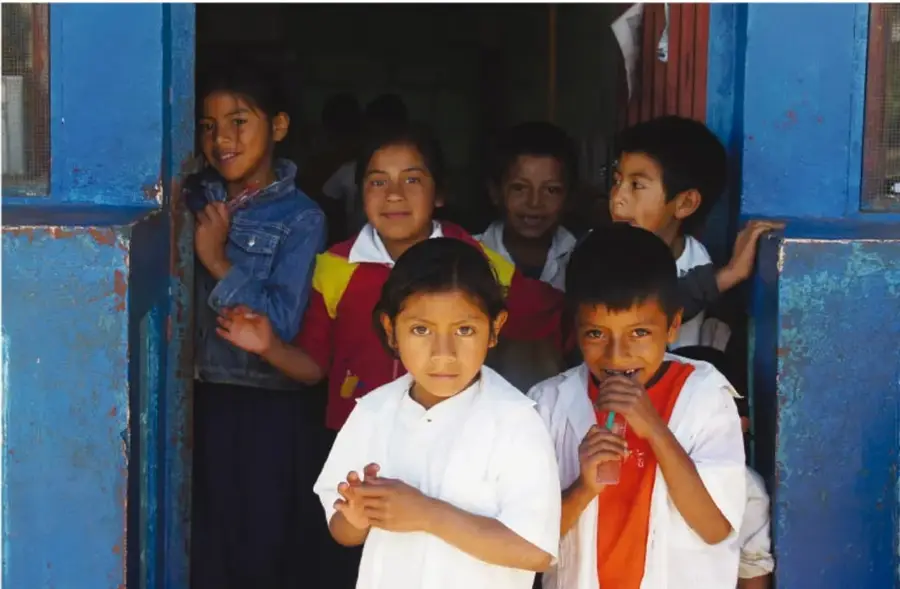 Fotografía de un grupo de niñas y niños de primaria asomados en la puerta azul de una escuela; sonríen y miran a la cámara. Representa la comunidad escolar y el trabajo colectivo.
