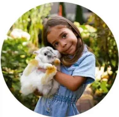 Fotografía circular de una niña sonriente que abraza con cariño a un conejo blanco entre vegetación.