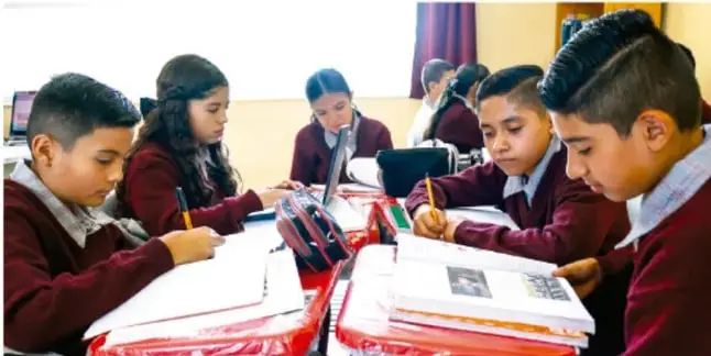 Fotografía de estudiantes de telesecundaria en uniforme trabajando con libros y tabletas alrededor de una mesa, desarrollando su proyecto académico en equipo.