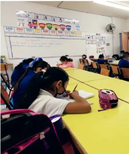 Fotografía de un salón de clases de primaria con estudiantes sentados en mesas amarillas, trabajando en sus cuadernos; al fondo hay un pizarrón con material didáctico.