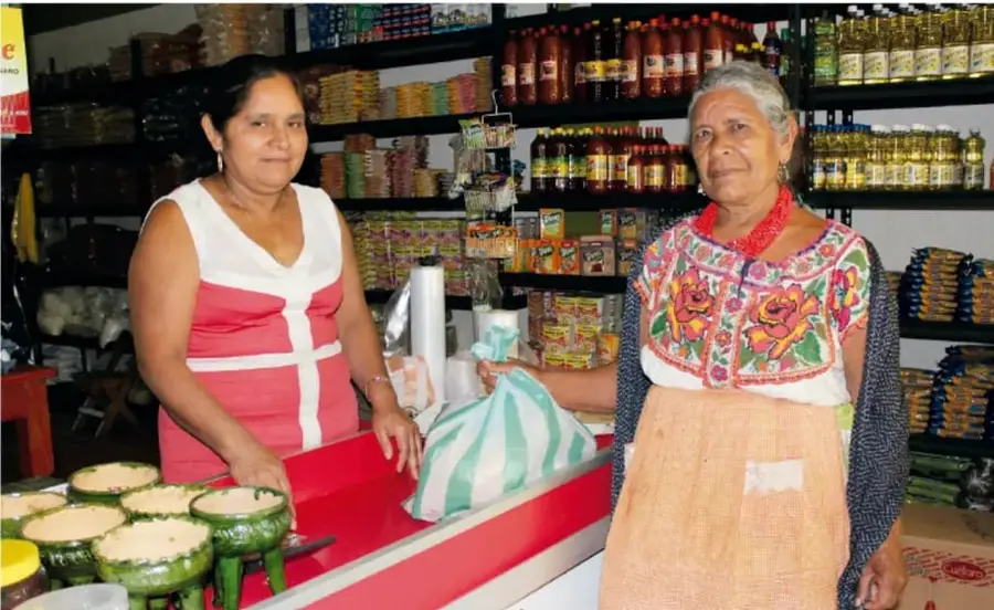 Fotografía de dos mujeres en una tienda de abarrotes. Una viste blusa rosa con blanco y la otra un huipil bordado con flores y delantal. Sobre el mostrador hay alimentos envueltos en hojas verdes y una bolsa de plástico verde y blanca; detrás se ven estantes con productos.