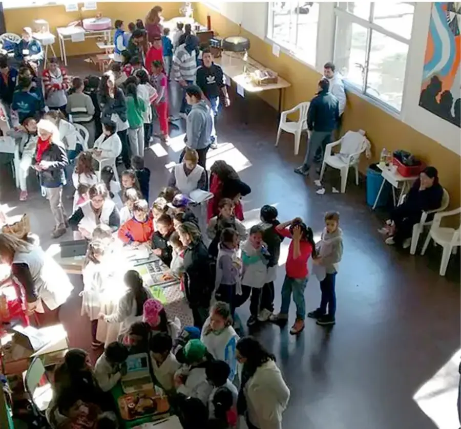 Fotografía de un trueque escolar en un salón amplio con mesas y puestos atendidos por niñas, niños y adultos; se observan productos, carteles y numerosos asistentes intercambiando.