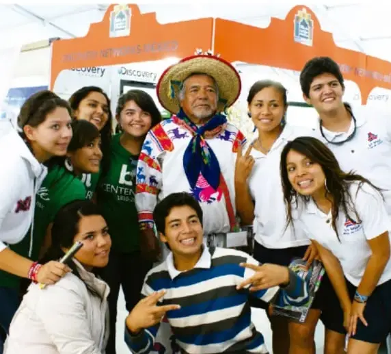 Fotografía de un grupo de jóvenes junto a un hombre adulto con sombrero, en un evento con stand del programa Discovery; ilustra la interacción intercultural.