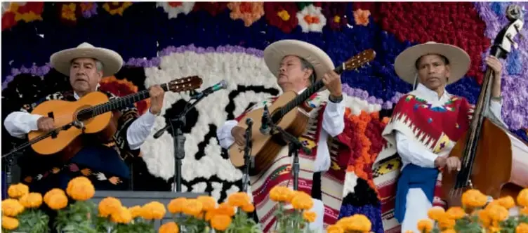 Fotografía de tres músicos con sombreros tradicionales interpretando instrumentos de cuerda (guitarra, vihuela y contrabajo) ante un fondo floral decorativo con flores de cempasúchil, posiblemente durante una festividad mexicana.