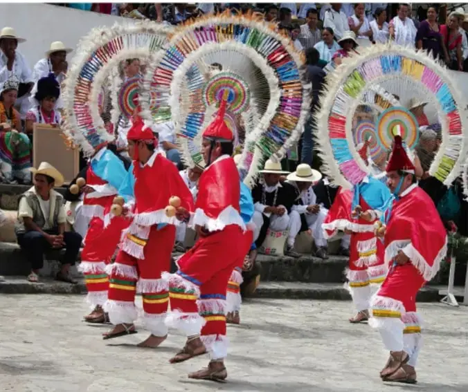 Fotografía de danzantes con vestimenta roja, sombreros con penachos de colores y máscaras, bailando en una plaza ante público sentado; ilustra el festejo o tradición investigada por los estudiantes.