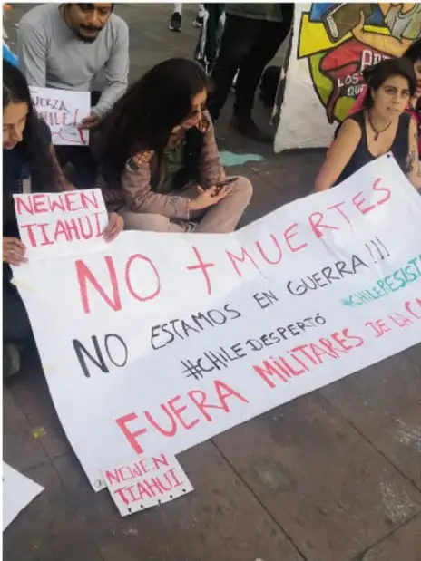 Fotografía de jóvenes en una manifestación pacífica sosteniendo carteles con mensajes como «NO + MUERTES», «NO ESTAMOS EN GUERRA», «FUERA MILITARES DE LA CALLE» y leyendas mapuche como «NEWEN TIAHUIL» y #CHILEDESPIERTO, #CHILERESISTE.