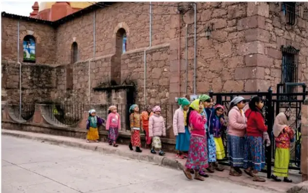 Fotografía de un grupo de mujeres y niñas ralámulis con vestimenta tradicional colorida, en fila frente a un portón junto a un edificio de piedra en Creel.