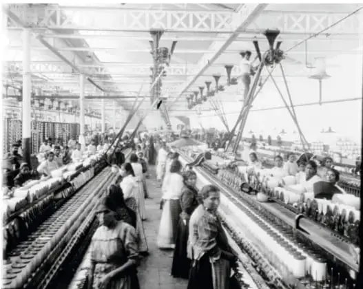 Fotografía en blanco y negro de una fábrica textil durante el Porfiriato. Se observan hileras de máquinas con husos y numerosos trabajadores, en su mayoría mujeres, operando el equipo en un amplio galpón industrial con estructura de hierro y techo a dos aguas.