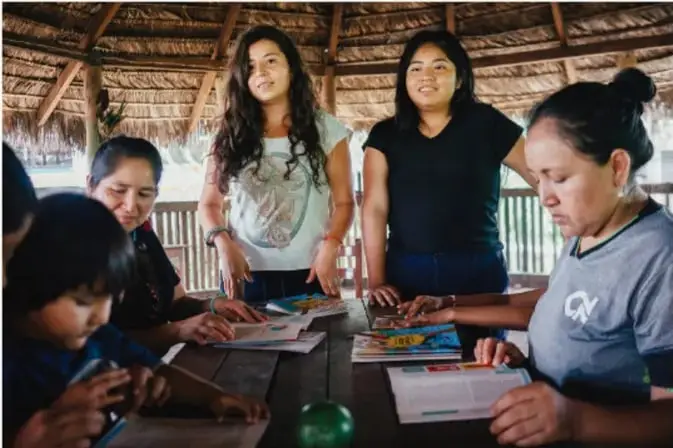 Fotografía de un grupo de personas de diversas edades reunidas alrededor de una mesa en un espacio techado de palma, trabajando con materiales impresos, probablemente en una actividad comunitaria o escolar.