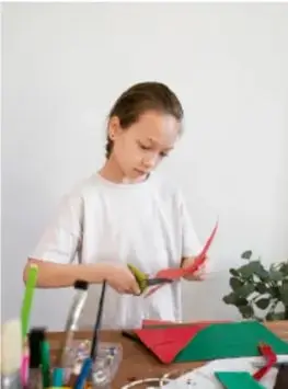 Niña construyendo un papalote con papel de color verde y rojo sobre una mesa.